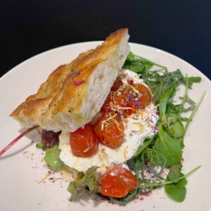 Frische Focaccia mit cremigem Käse, geschmorten Kirschtomaten und buntem Blüten-Topping auf einem Bett aus knackigem Rucola auf einem weißen Teller.