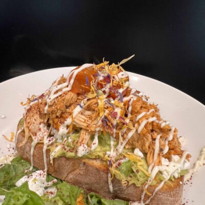 Geröstetes Brot mit Avocado, gezupftem Hähnchen, weißer Sauce und essbaren Blüten auf einem weißen Teller, serviert mit frischem Salat als Beilage.