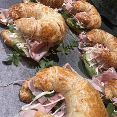 Mehrere goldbraune, knusprige Croissants, gefüllt mit feinem Schinken, Käsescheiben, frischem Rucola und einer cremigen Mayonnaise-Garnitur.