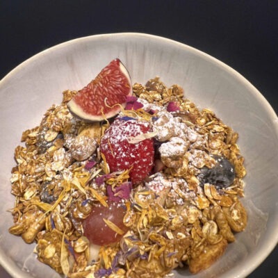 Eine Schale mit knusprigem Granola, garniert mit frischer Feige, Erdbeere, Weintrauben und essbaren Blüten, leicht mit Puderzucker bestäubt.