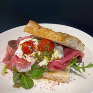 Ein knuspriges Ciabatta-Sandwich mit feinem Schinken, cremiger Burrata, Kirschtomaten, frischem Pesto und Salat auf einem weißen Teller serviert.