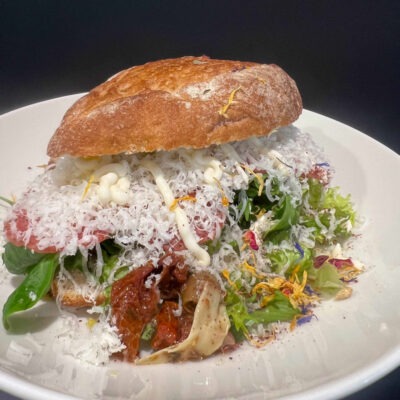 Ein rustikales Sandwich mit Salami, getrockneten Tomaten, frischem Salat, essbaren Blüten und einer großzügigen Schicht geriebenem Käse auf weißem Teller.