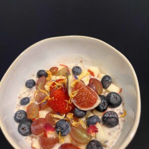 Eine cremige Bowl mit frischen Blaubeeren, Erdbeeren, Feigenstücken und Trauben, garniert mit essbaren Blüten in einer hellen Keramikschale serviert.